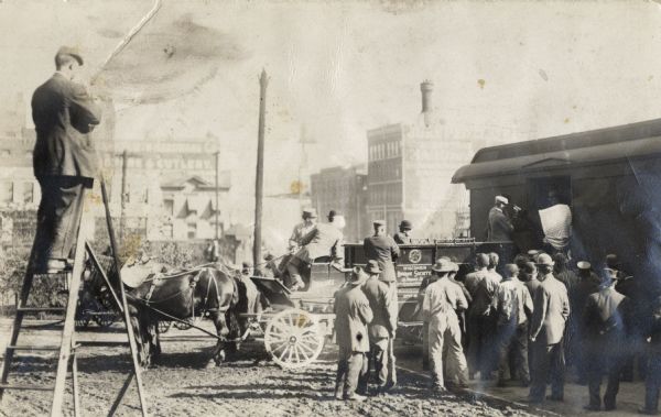 A group of men are gathered around a horse-drawn cart that is pulled up next to a railroad car.  The cart has "Wisconsin Humane Society" written on its side. From the train, two men are leading a cow from the train to the cart. On the left is a man standing on a ladder watching (or possibly photographing) the event.