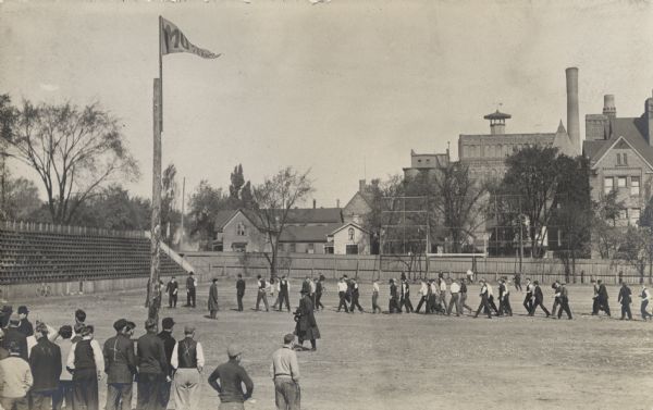 Elevated view of a line of young men walking across an athletic field. In the left corner is a group of onlookers, some of which have markings on the back of their jackets. A flag with "MU 2nd Year" is flying from a pole.