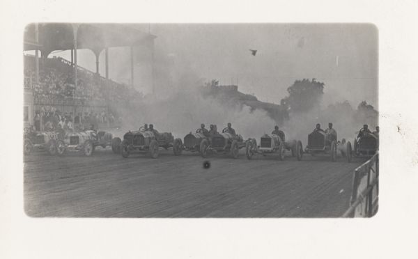 Early twentieth-century race cars lined up for the beginning of a race. heavy smoke is behind the cars. The grandstand on the left is filled with spectators.