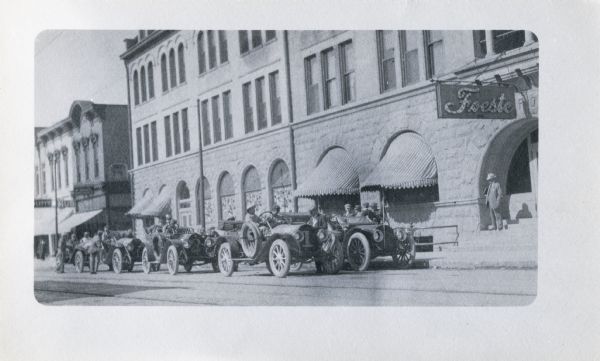 Men with several early twentieth-century race cars are parked along a city street. Numbers are painted on the front of the cars. One man is standing on steps near a doorway of a large building. The sign above him reads "Foeste".