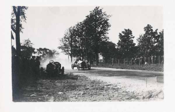 Race cars on a country road, with car #29 off the road and missing its driver. The #27 car is just about to pass, with a dust trail behind it.  Spectators are along both sides of the road.
