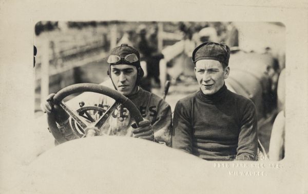 Driver Spencer Washart on left, with John Genter (the mechanic) sitting next to him. Washart has 'Mercedes" written across his shirt. Another car is behind them, with people on a fence in the background.