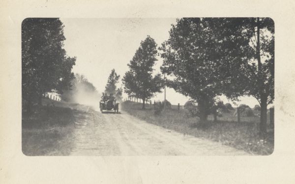 Four men in a car with the number four on the front is driving up a dirt road surrounded by fields. Hay bales are in the field on the right.