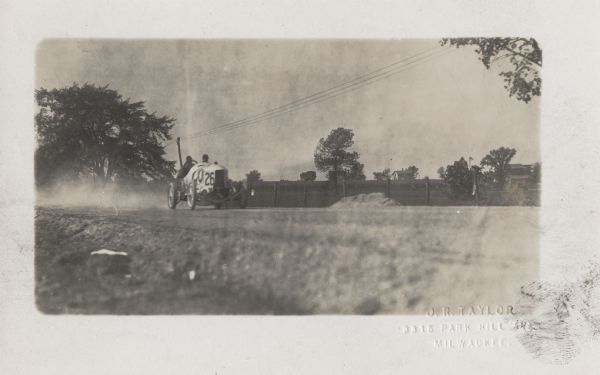 Race car #26, possibly driven by Spencer Wishart, driving down a country road during the Vanderbilt Cup Race. Two houses are on the right behind fences and fields.