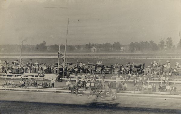 Elevated view of a race car on the track moving past a stationary vehicle in the foreground. Behind a second fence is a large crowd of people observing the races, near a row of parked automobiles. Beyond the track is a residential area.