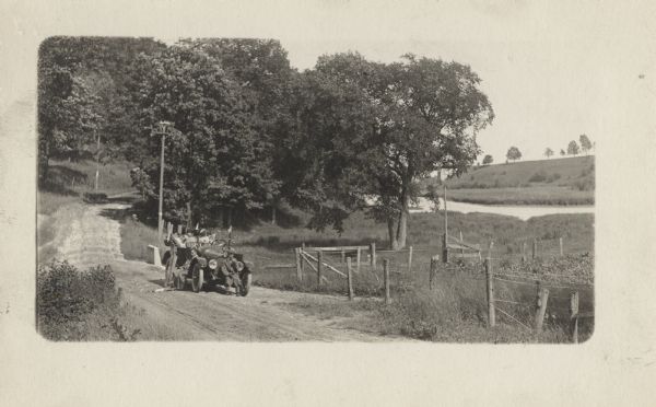 The Milwaukee Journal car on the side of the road in the countryside. Two women are sitting in the car, and two men are sitting on the side and front of the car. In the right background is a lake.