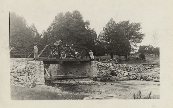 View from shoreline towards the Milwaukee Journal Pathfinder car parked on a bridge. Under the bridge is a stream with a small waterfall. Two women are standing on either side of the bridge, and a man is sitting on the railing of the bridge.