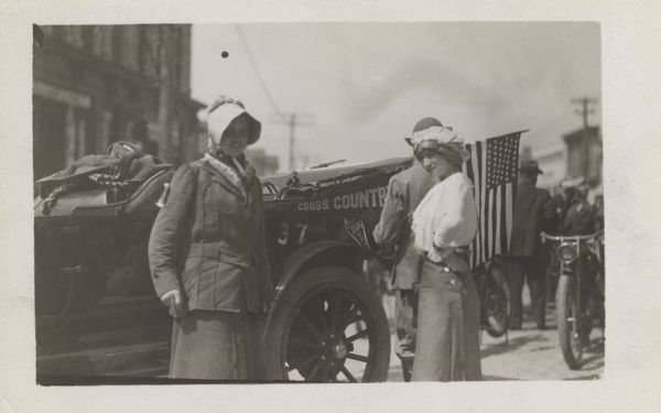Two women and a man are standing in front of the Pathfinder car which has a flag on the back.  The car also has "cross country" written on the side, above the number 37. There are people and motorcycles in the background.