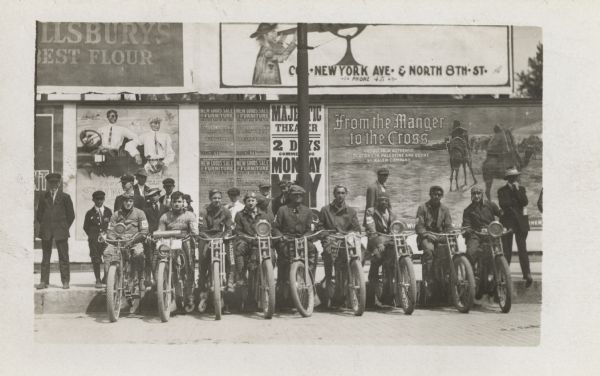 Nine men are lined up along a curb sitting on their motorcycles. Men and boys are on the sidewalk behind them, wtih advertisements filling the wall behind the group.