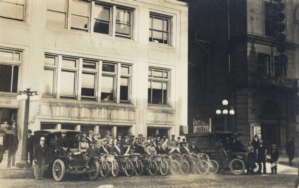 Eleven motorcyclists wearing sashes are standing in between two news service cars in front of the <i>Milwaukee Journal</i> building. Another row of men are standing behind them on the sidewalk, and various other men are around the automobiles parked on the left and right. A boy us standing on the right with a sign that reads: "22 only". A large poster for the Sunday edition of the <i> Milwaukee Journal</i> is in the alley.