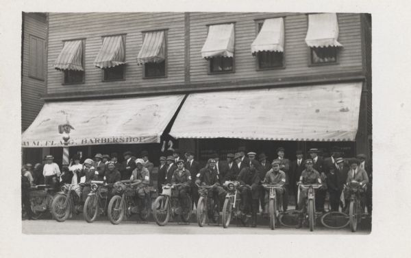 A group of motorcyclists, both male and female, are lined up in front of a sidewalk, with a group of people standing behind them in front of an awning for a barbershop.
