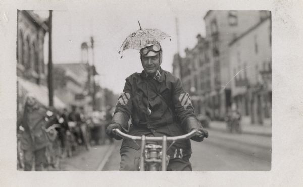 A man on his motorcycle in the street is posing with a small umbrella attached to his helmet. A watch also hangs from the front of his shirt.