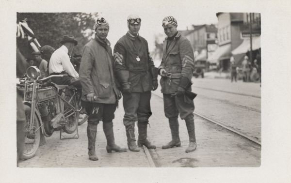 Three men are posing in the street to the right of a Harley-Davidson motorcycle with a group of men in the background. The man in the middle of the group has a watch hanging from his shirt. Two of the men are wearing striped caps.