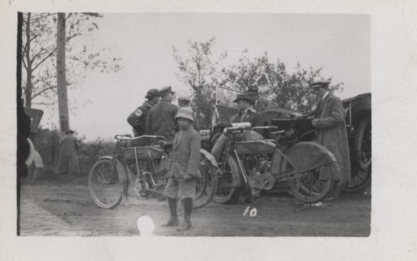 Several men are standing and talking by a car, with Harley Davidson motorcycles in front of them. A child is standing in the foreground.