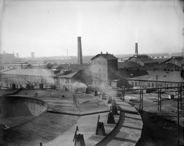 Located in West Milwaukee. View from atop a building of a large building located in the center, next to a smokestack.  In the foreground is part of a rounded building (railroad roundhouse), with railroad tracks in the lower right shadows.