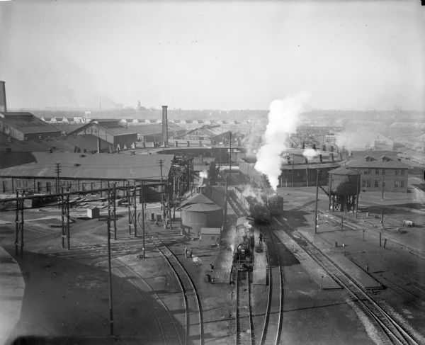 West Milwaukee. Elevated view of tracks, trains, and buildings.  A train roundhouse is located in the background.
