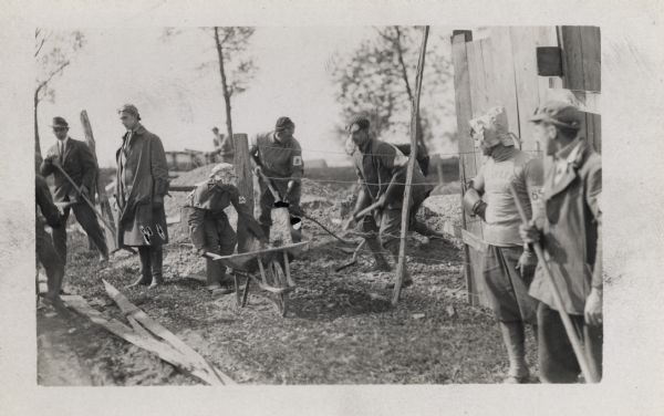 Several people, most wearing goggles and hats, are digging and moving dirt. Three men are behind a wire fence. On the left is a man in a suit. The rest of the men are wearing traveling clothes.