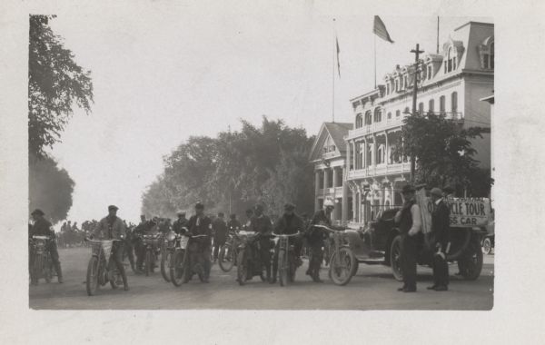 In the middle of a road, a large group of motorcyclists are preparing to ride. On the right are two men standing next to a car which has a banner advertising for the <i>Milwaukee Journal</i> Motorcycle Tour.