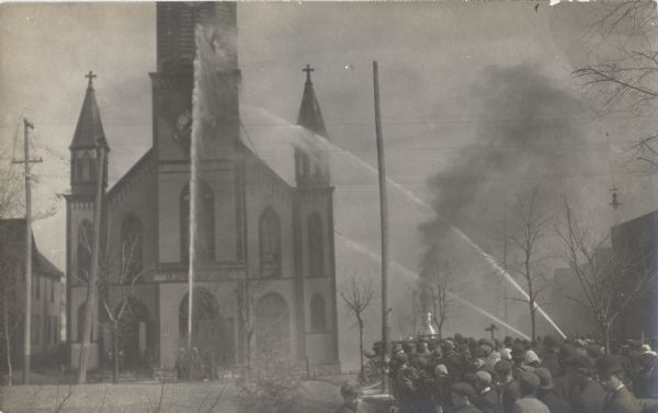 A crowd is gathered on the street as jets of water are being directed by fire fighters towards a fire in a church.