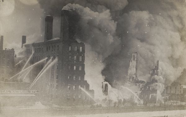 A large building has partially collapsed from a fire. Fire fighters, some of them standing on the roof of a building next door, aim large streams of water with fire hoses. Clouds of billowing smoke stream out of the chimneys on the roof.