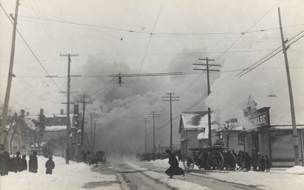 A large cloud of smoke blankets the end of the road, and snow covers the ground. Pedestrians are the sidewalks. A woman is running across the street, and a fire engine is parked in front of a hardware store on the right.