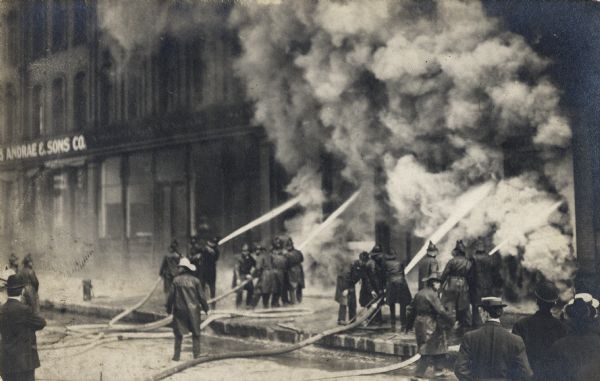 Groups of fire fighters work to put out a fire in winter. Smoke is billowing out the windows and covering the facade. A building on the far left has a sign for Andrae & Sons Co.