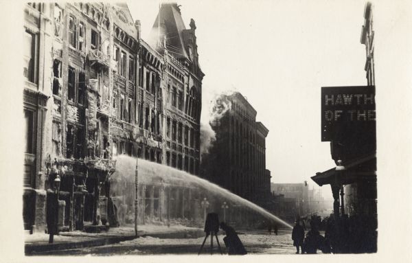 Fire fighters work to put out a fire in a four-story building on a city block. In front of a fire hose spraying water, a photographer is kneeling in front of a large format camera, preparing for a shot.