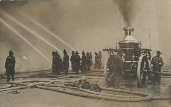 A small group of fire fighters fight an unseen fire with three fire hoses. Smoke obscures the fire, and on the right, a fire engine has smoke billowing from its steam engine.