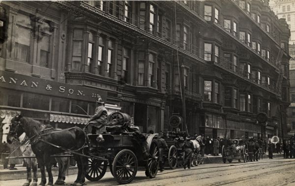 A fire fighter driving a horse-drawn fire engine is parked on the street. A group of people are looking up toward a building where a rope is coming down from the upper floors. Fire hoses are lying in the street. Signs for "Turkish Baths" and "Capt.' Lewis" are on some of the buildings.