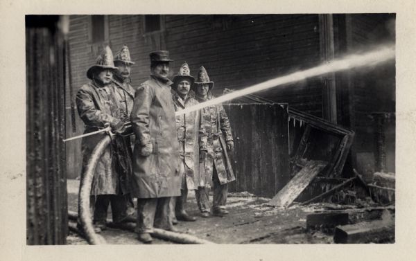 A small group of four fighters and one of their bosses are standing amid burned remnants from a fire. One man is holding a fire hose that is spraying water. the water has frozen on their jackets and icicles have formed on their hats.