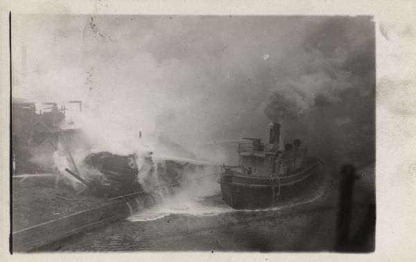 A fire boat is next to a dock with burning objects. Fire fighters on the boat are using high-powered hoses to put out a fire. Smoke and steam fill the background.