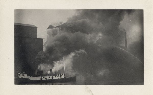 View across water towards fire fighters in a boat on the river putting out a fire in a large building. Water sprays from fire hoses from two directions, with smoke obscuring the burning building.