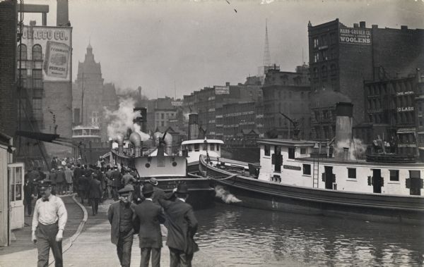 Boats used for fighting fires are docked near a walkway. On the left the walkway is full of men, some gathered near the boats and a fire hose stretched out on the ground.
