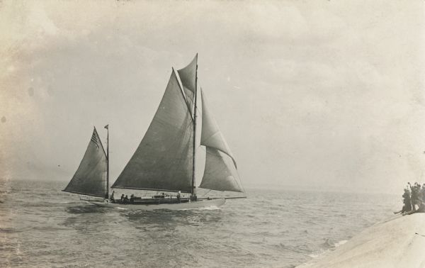 A group of people on a sailboat sailing towards a group of people who are waving from a concrete embankment.