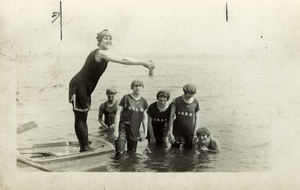 A woman is standing in a rowboat in a diving position teaching four women in a lake how to dive. The rest of the women are in shirts printed with S.S.B.H. printed on them and are standing in the water. They wear bathing caps and long pants. A man in a hat is standing in the water in the background.