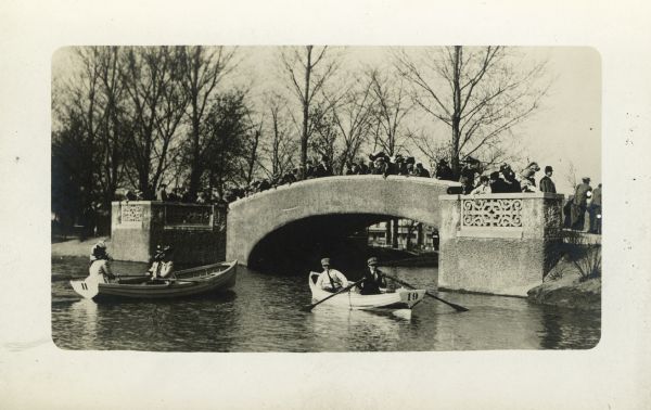 Rowboat race with two boats about to go under a bridge. Both of the boats have numbers on the back. Onlookers are standing on the bridge watching the participants.