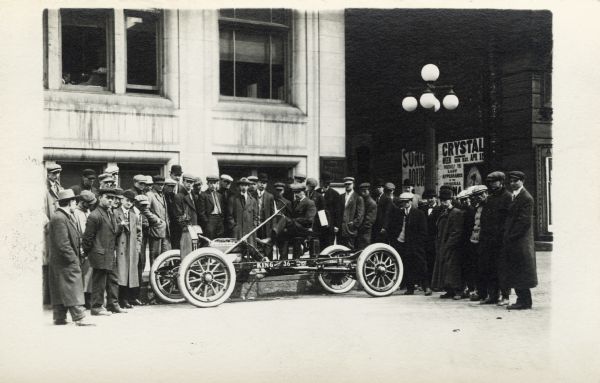 Crowd of men gathered around a man sitting in an early automobile with exposed wheels, engine, and steering wheel. The side of the automobile reads: "King Silent 36". The group is probably gathered in front of the <i>Milwaukee Journal</i> building. There is an alley with posters on the right side of the building.