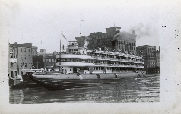 Passenger ship "Christopher Columbus," which is full of people on multiple decks, along the shore of the city of Milwaukee. Buildings and a second ship are in the background.