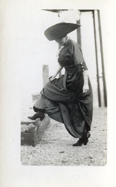A woman wearing a large feathered hat is standing and looking down at her shoe which she has rested on a step. She is wearing a split skirt.