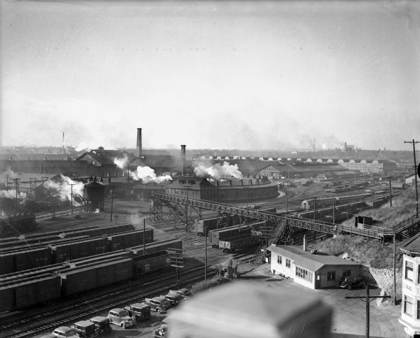 Located in West Milwaukee.  Trains, cars, tracks, and railroad buildings. The view is taken from the top of a building.
