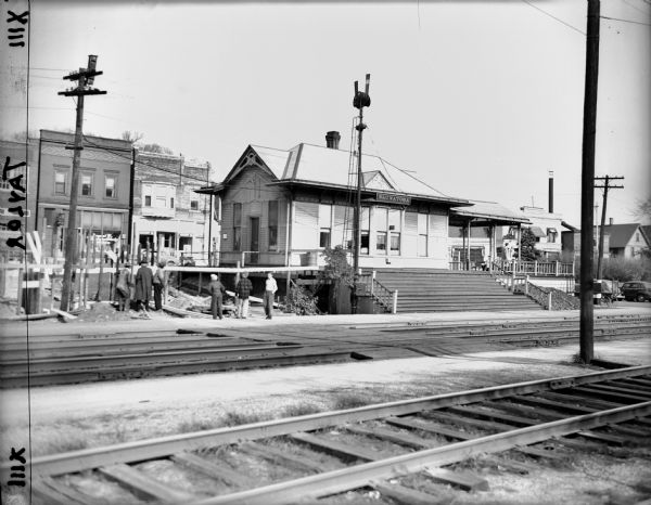 View across railroads tracks towards the Chicago, Milwaukee & St. Paul Railroad station. A number of children and adults are standing around the station. Storefronts line the street in the background.