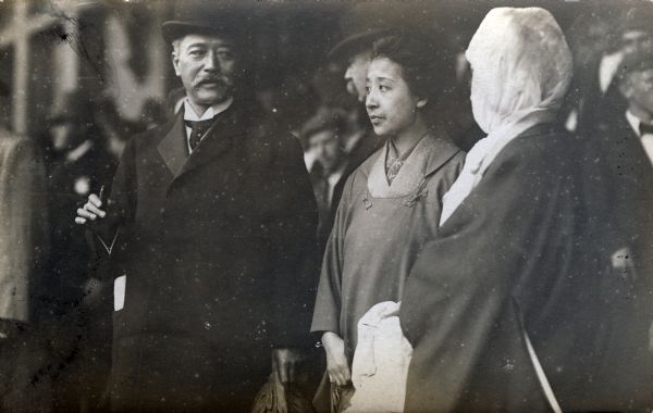 Asian dignitaries, including a man and two women, are standing amidst a crowd, perhaps at a railroad station.