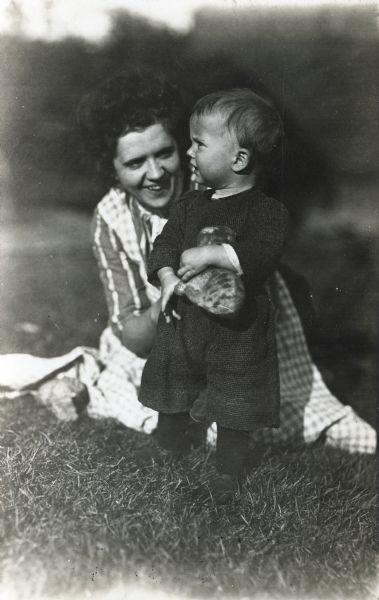 Alma Reinhardt Taylor playing on the grass with her son, Frederick or Robert. The child is holding a stuffed animal toy.