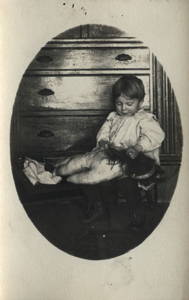 A child is sitting on a small chair in front of a dresser holding a stuffed toy. A doll is sitting on a bench nearby.