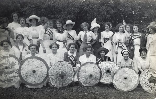Women posing for a group portrait at the Wisconsin Telephone Company Employees' Picnic. Women in the front row are holding open parasols in front of them, and other women are holding or wearing pennants which read: "Grand".