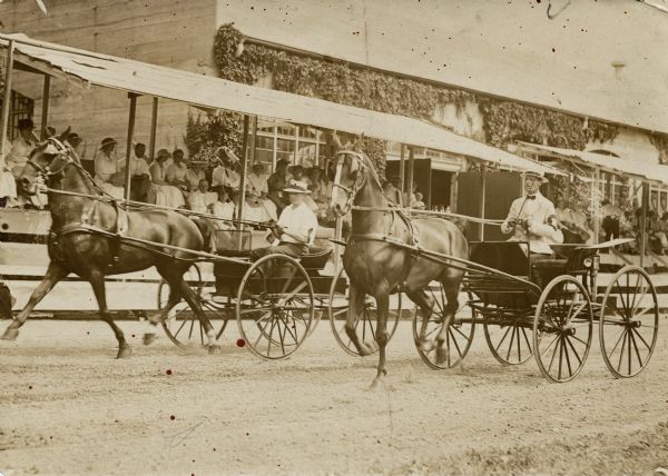 Two men are racing small horse-drawn buggies on a dirt track at an exhibition at the Pabst Farms. The man on the left is identified as Fred Pabst, Milwaukee brewer. Groups of people are watching from the stands. Behind the stands is a vine-covered building.