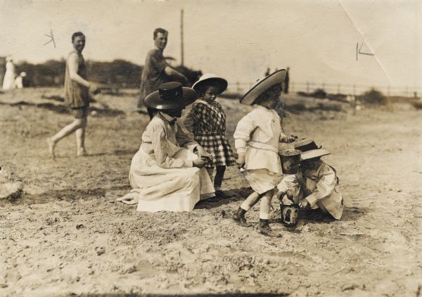 A woman and three children, all wearing hats, dresses, and shorts, are at the beach, playing in the sand. Two of the children have toy buckets. Behind them, two men in bathing suits are walking by smiling.
