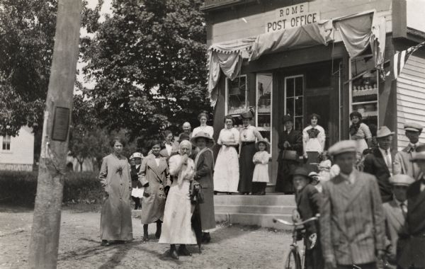 A crowd of people looking toward the camera has gathered in front of the Rome Post Office that has a folded-up awning and a flag over the entrance. A pathway to the entrance has formed between the men and boys on the right, and the women and girls on the left. An older woman in white stands in front of the other women with her hand to her mouth. A boy on the right has a bicycle.
