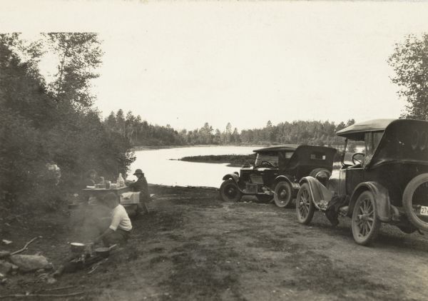 Cars are parked on the right near the water. Two people are sitting at a picnic table near the shoreline, and a third person is holding a pot over a campfire in the foreground.