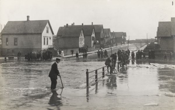 A river has overflown its banks and is flooding a road on a bridge.  Some men are on the bridge are using sticks to move ice floes. Crowds of people are watching from the road in the background, which is lined on both sides with houses.